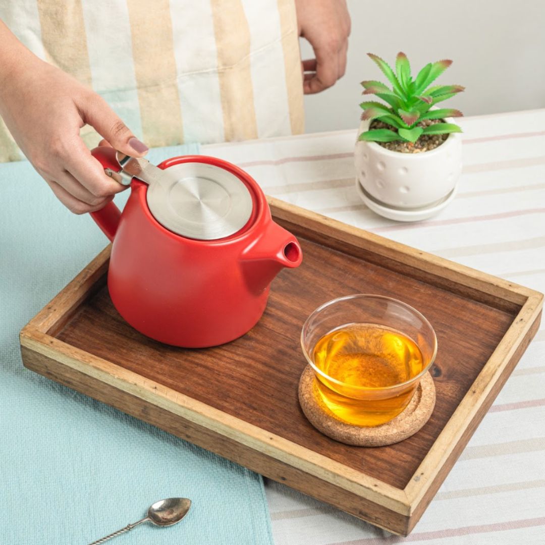 A person lifting the stainless steel lid of a red ceramic teapot placed on a wooden tray alongside a glass cup of tea on a cork coaster.
