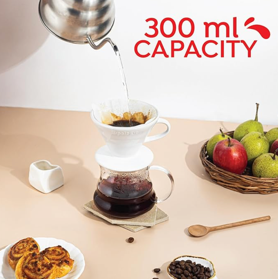 A coffee pour-over setup featuring a white dripper atop a glass carafe, with hot water being poured from a gooseneck kettle. The text '300 ml Capacity' appears in the background, surrounded by a fruit basket, pastries, and coffee beans