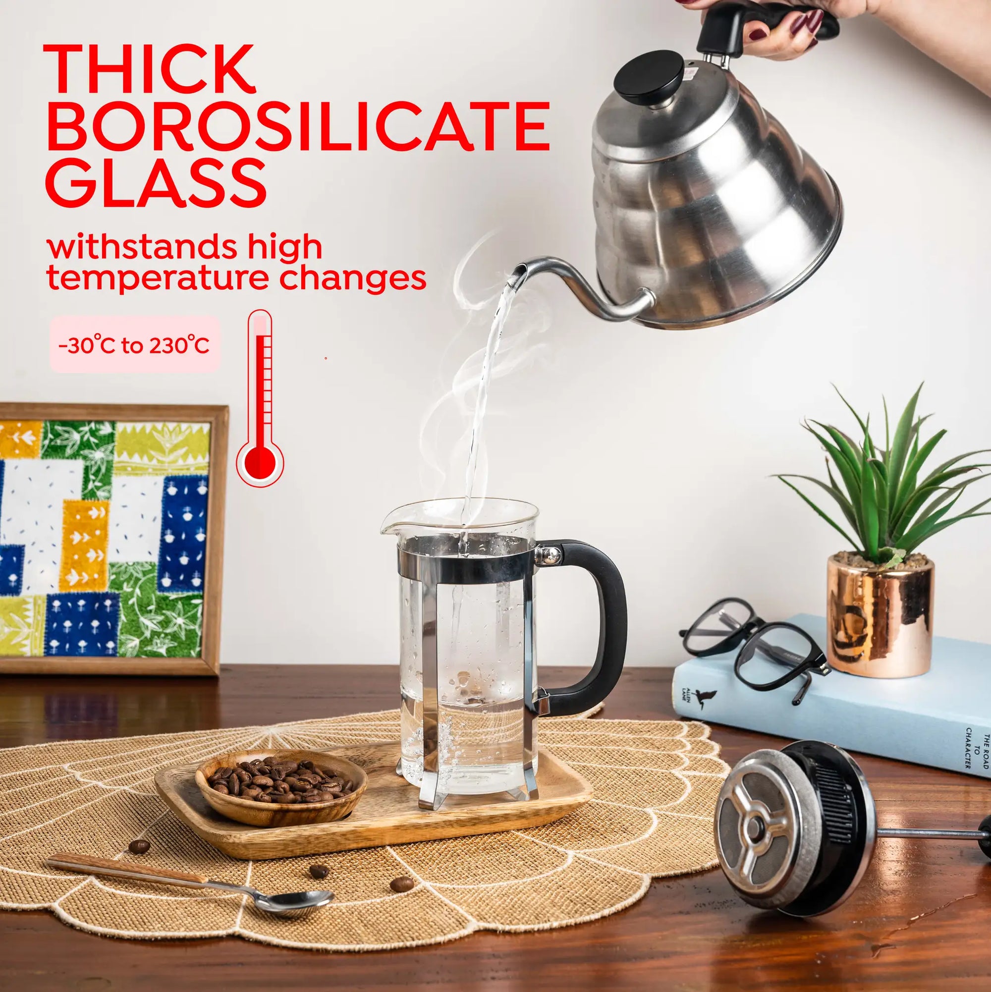 A clear thick borosilicate glass French press with a black handle and lid is shown on a wooden table with a metal kettle pouring hot water into it. Text on the left reads 'THICK BOROSILICATE GLASS' and 'withstands high temperature changes'.