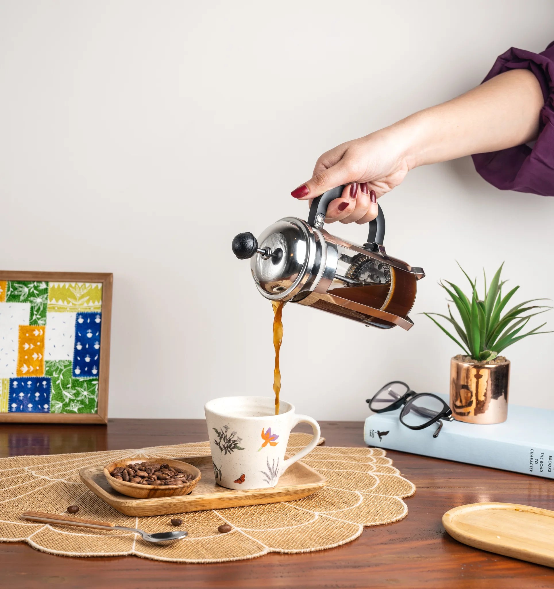 A person's hand pours hot coffee from a stainless steel French press with amber-brown liquid into a white ceramic mug with a floral pattern, set on a wooden tray on a table runner. The background features a small potted plant, glasses, a book, and a colorful framed print against a plain wall.