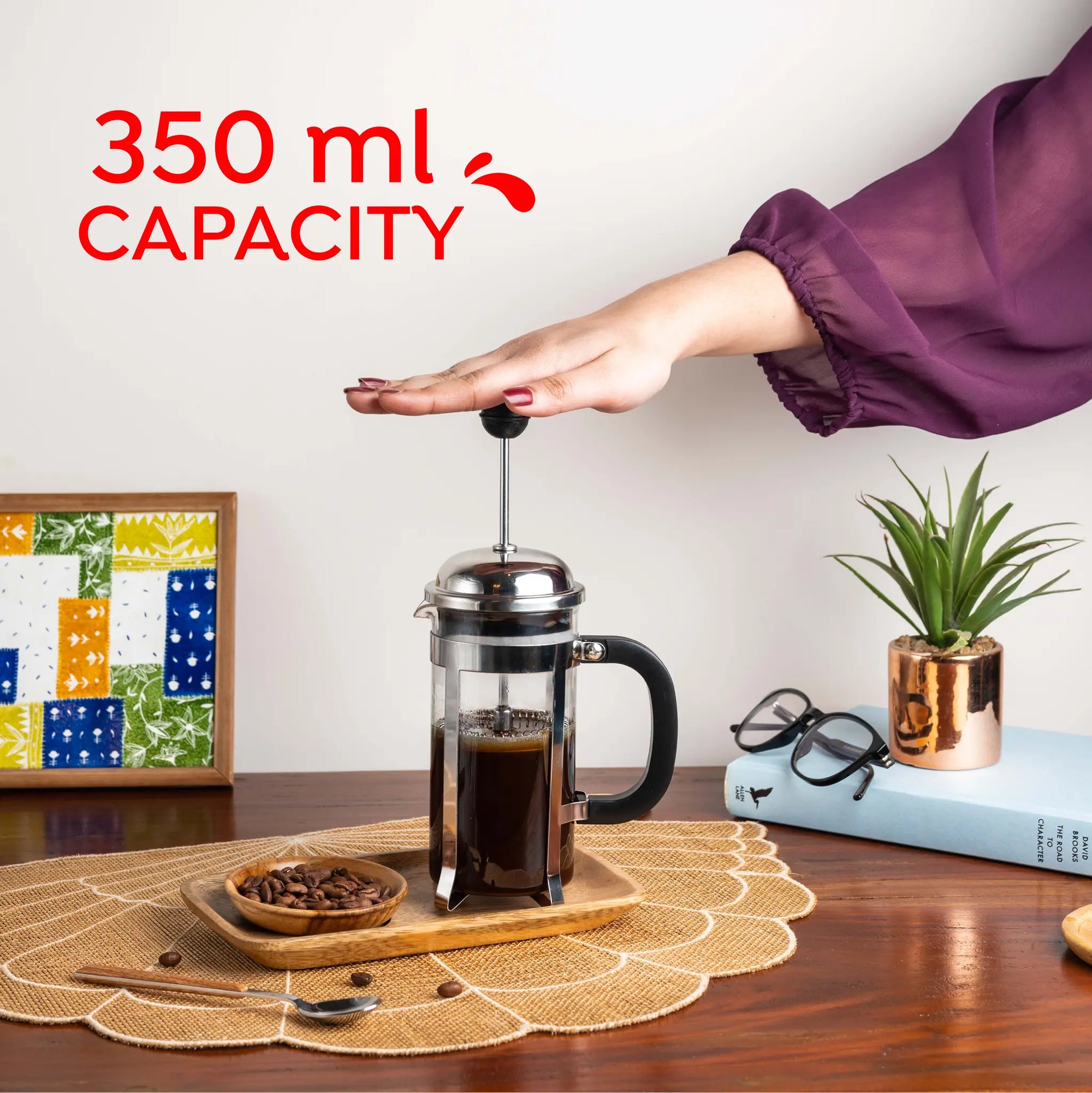 A hand presses the plunger of a 350 ml French press coffee maker on a wooden table, with coffee inside. The scene includes a small plant in a metallic pot, a book, glasses, a bowl of coffee beans, and a placemat.