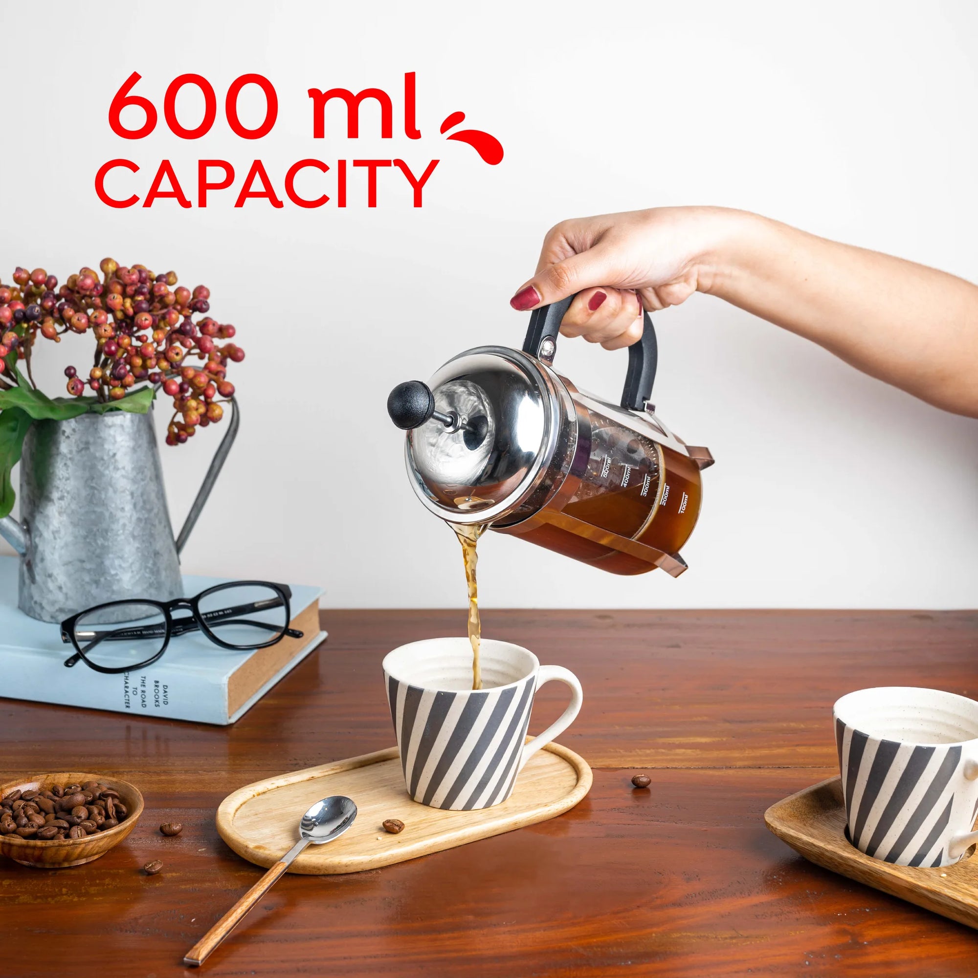 A hand pouring coffee from a stainless steel French press into a striped ceramic cup on a wooden table, with text overlay reading '600 ml CAPACITY'.