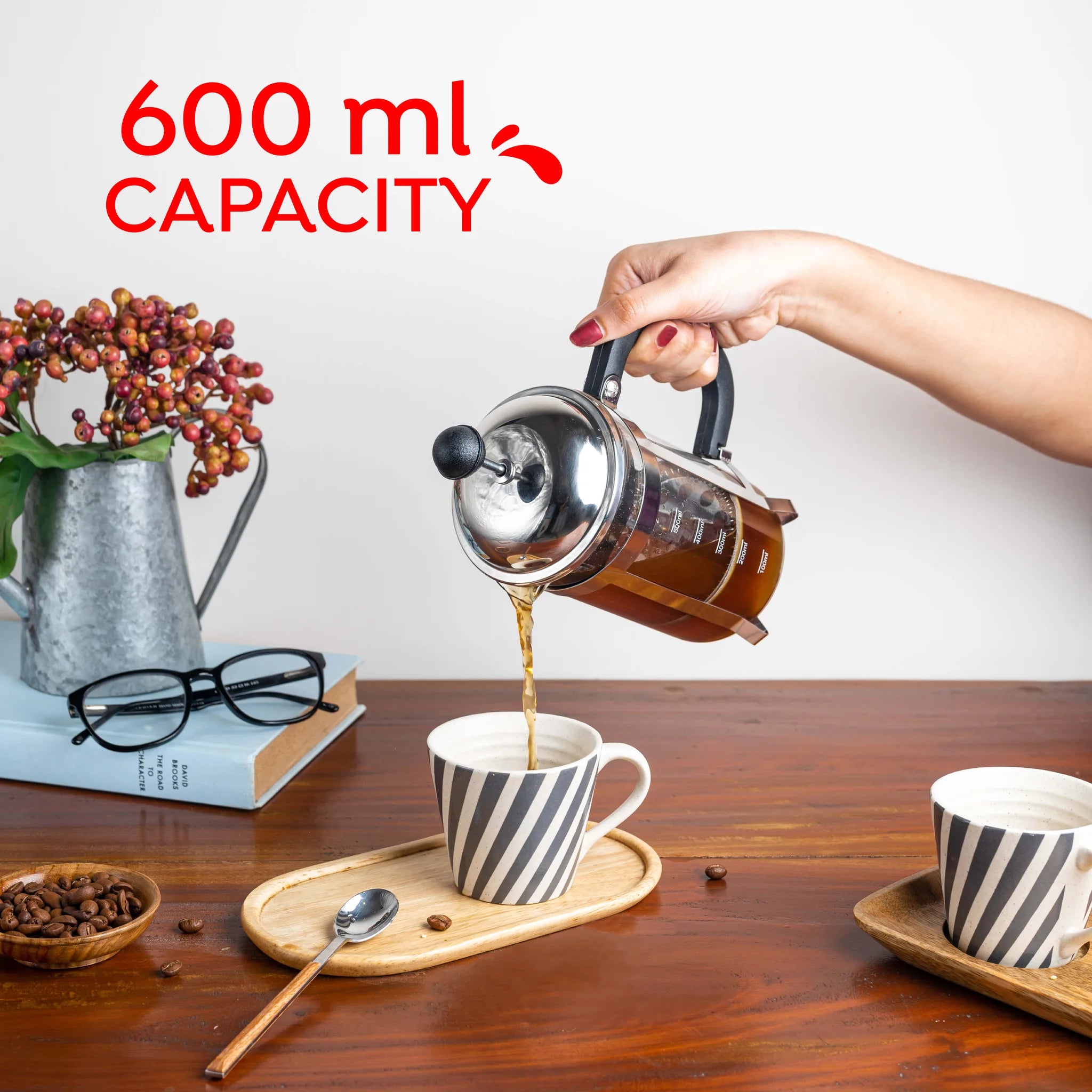 A hand pouring coffee from a stainless steel French press into a striped ceramic cup on a wooden table, with text overlay reading '600 ml CAPACITY'.