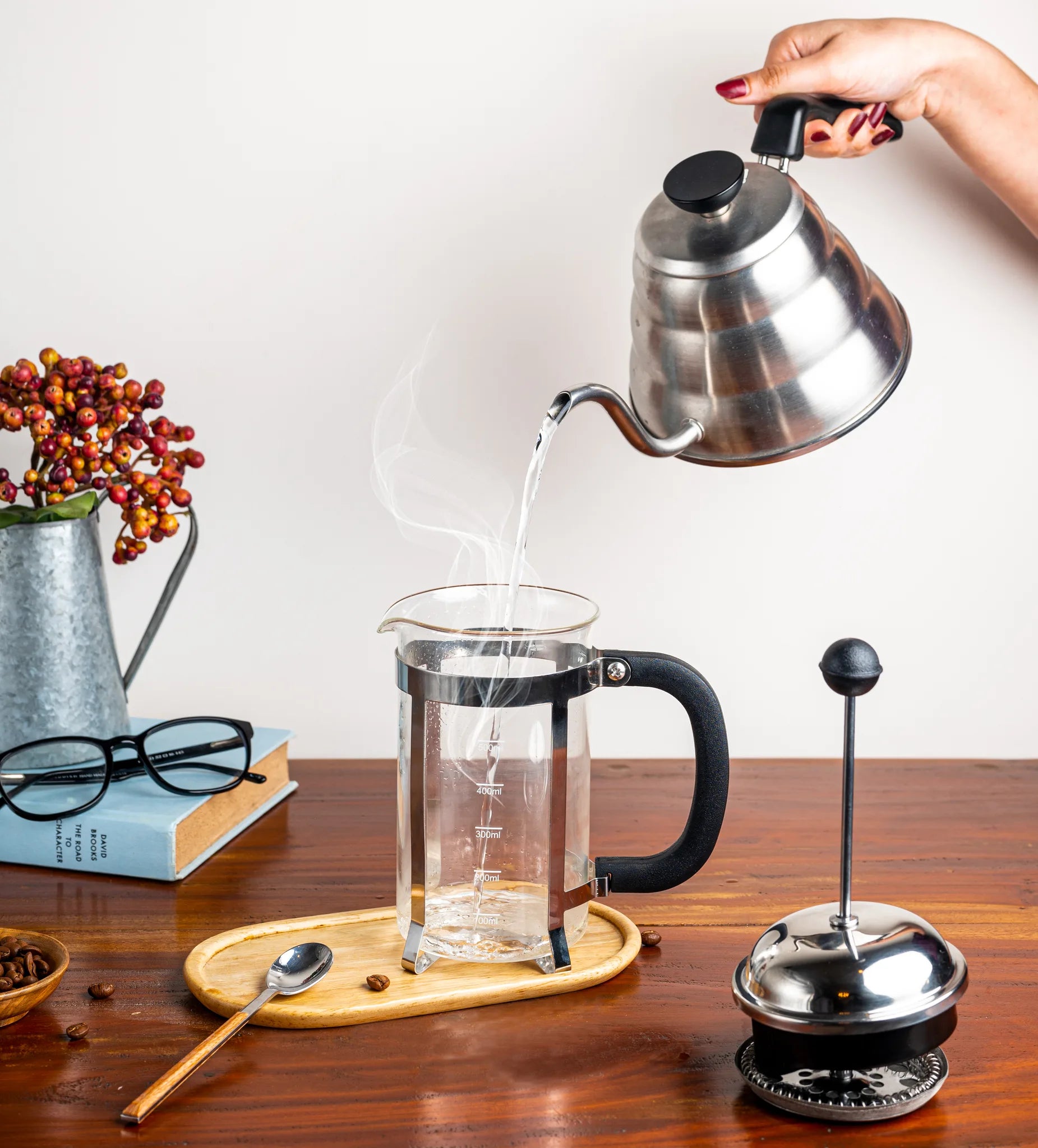 A glass French press with a black handle sits on a wooden table while a hand pours hot water from a stainless steel kettle into it. A metal coffee press tool and a small spoon rest beside the press, with books, glasses, and a decorative vase in the background.