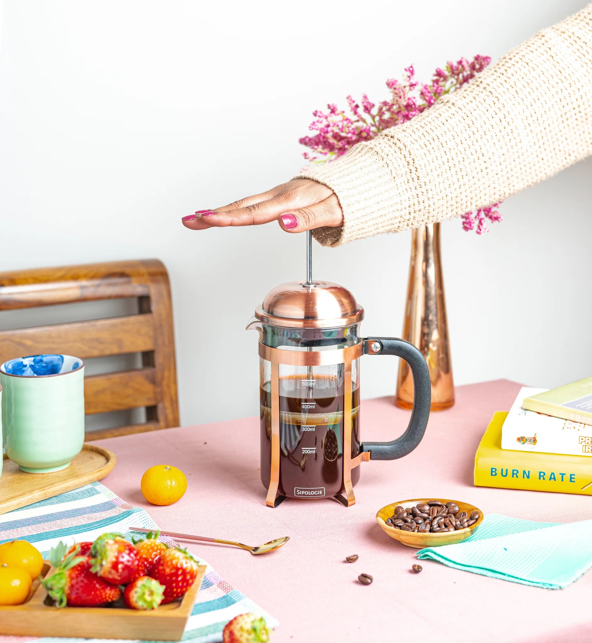 A copper-toned French press coffee maker with a black handle sits on a pink tabletop, with a person's hand pressing the plunger. The scene includes strawberries, coffee beans, a mug, books, and flowers in a styled indoor setting.