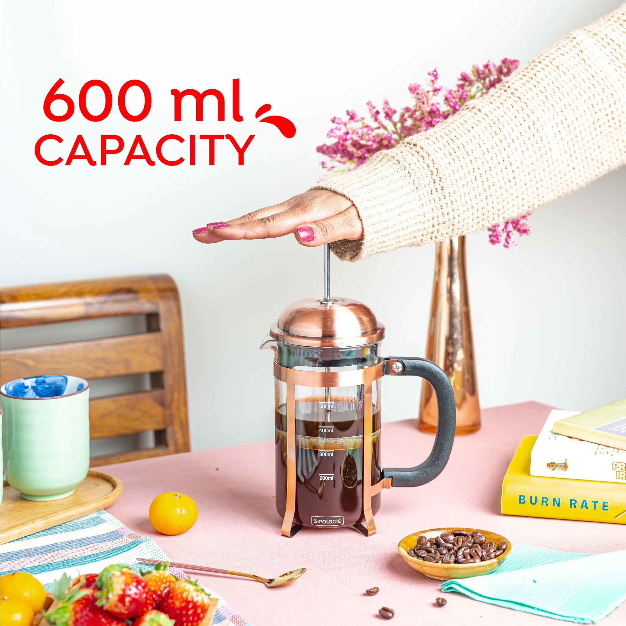 A French press coffee maker with a copper-toned metal body and black handle sits on a pink tabletop. A person's hand is pressing the plunger, and the text '600 ml CAPACITY' appears in red at the top left.