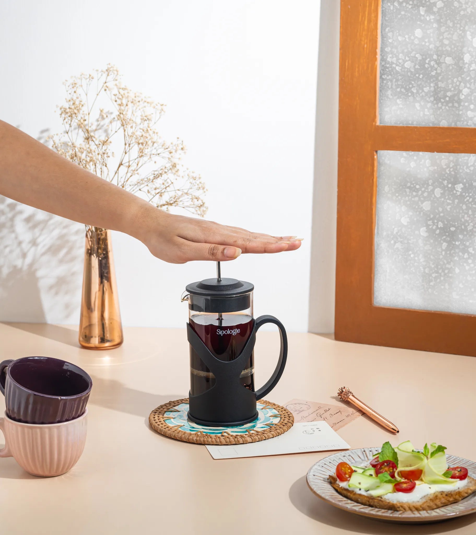 A black French press coffee maker with a handle sits on a woven coaster on a beige tabletop. A person's hand is pressing the plunger.