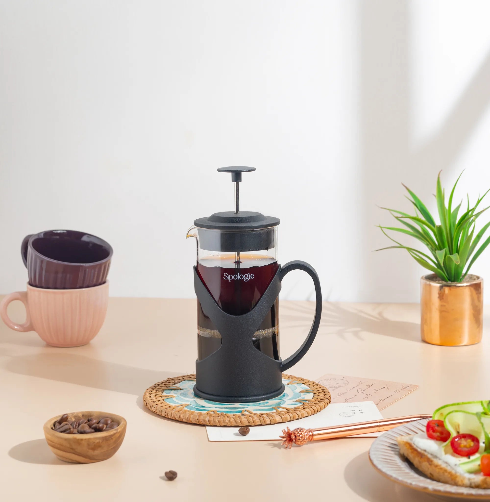 A black and clear glass French press coffee maker with the brand name 'Spoklinge' visible on the front, styled on a woven coaster on a beige tabletop with coffee cups, a small plant, and a slice of toast in a bright, clean setting.