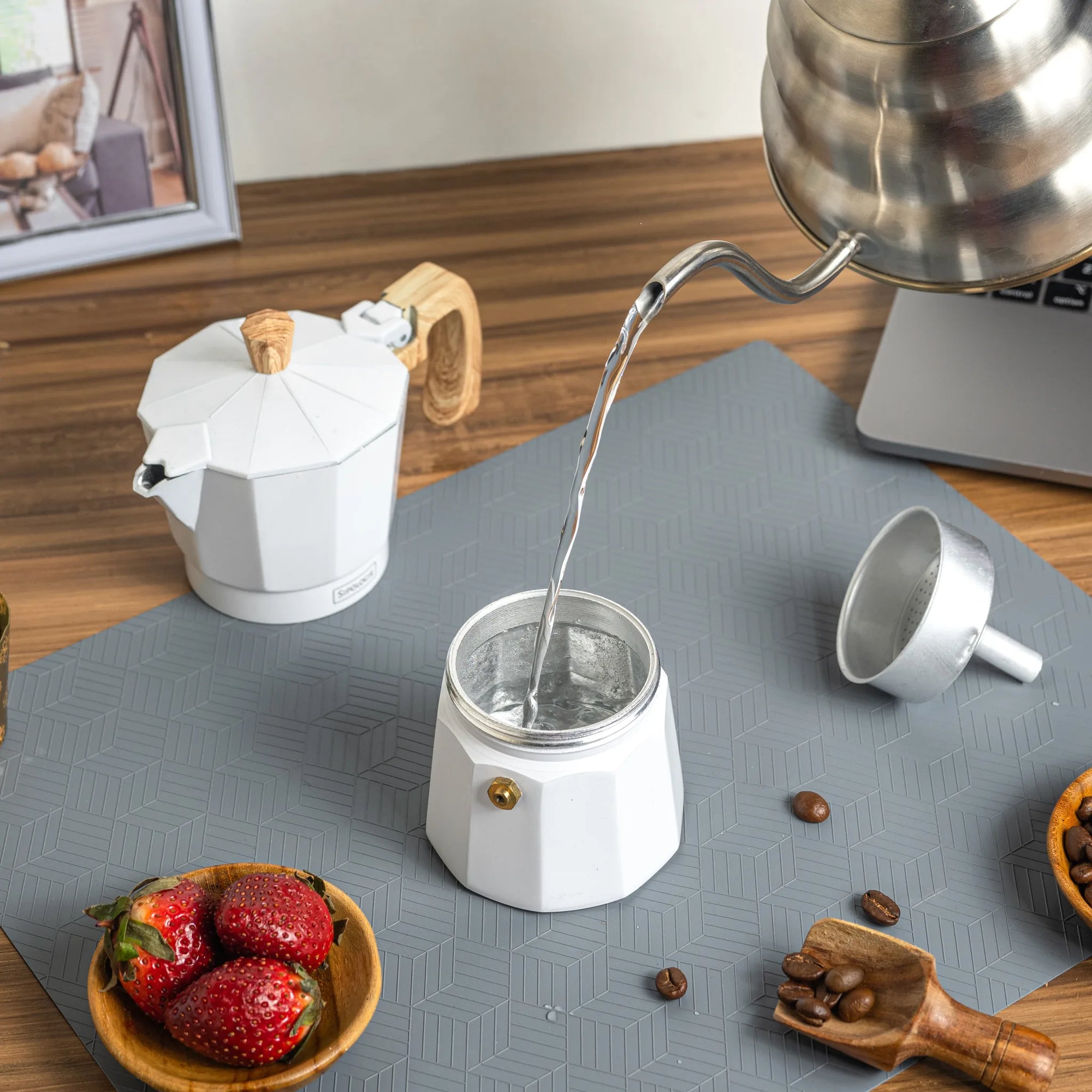 A white stovetop moka pot with a wooden handle sits on a gray mat while water is poured from a stainless steel kettle into the pot. A small metal cup, coffee beans, strawberries, and a wooden scoop are arranged around it on a tabletop.