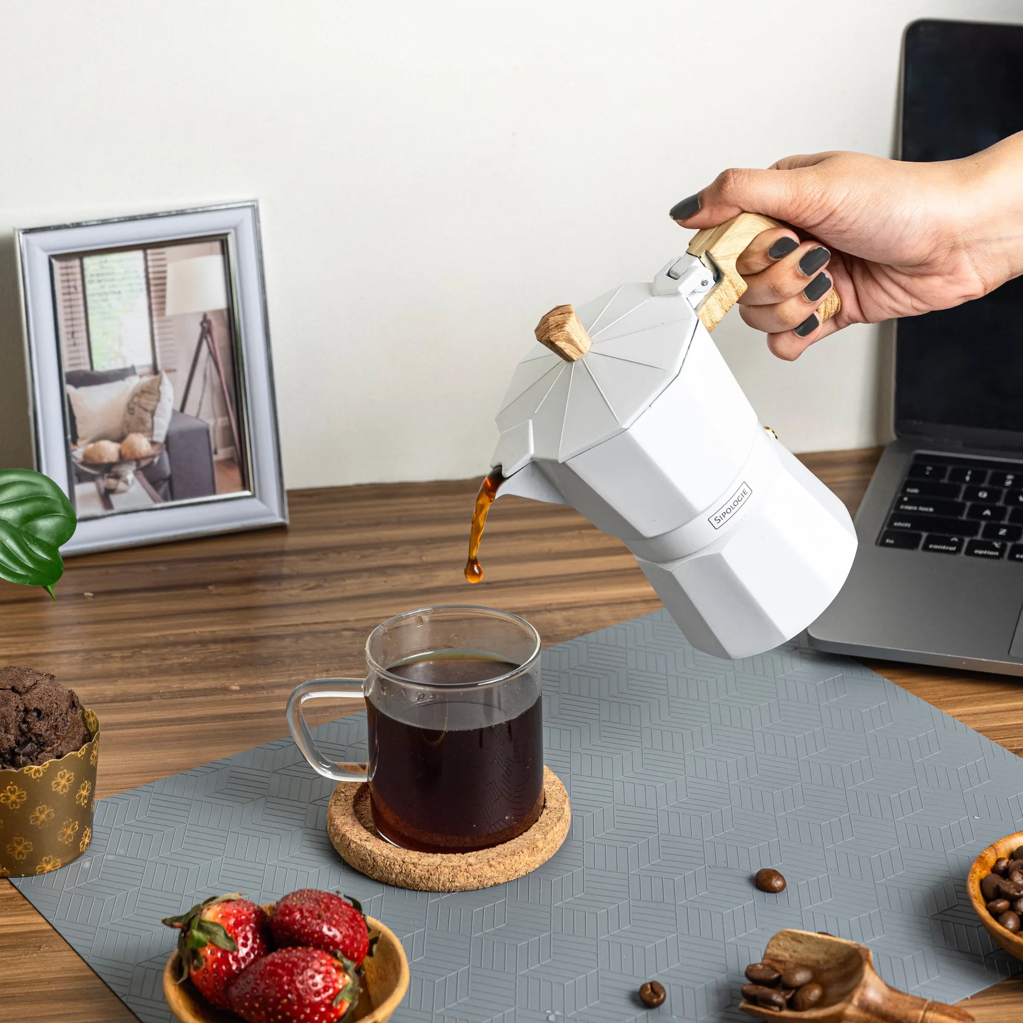 A white stovetop moka pot with a light wood handle is being used by a hand to pour coffee into a clear glass mug on a coaster, set on a gray mat on a wooden desk with a laptop, strawberries, and a framed photo in the background.