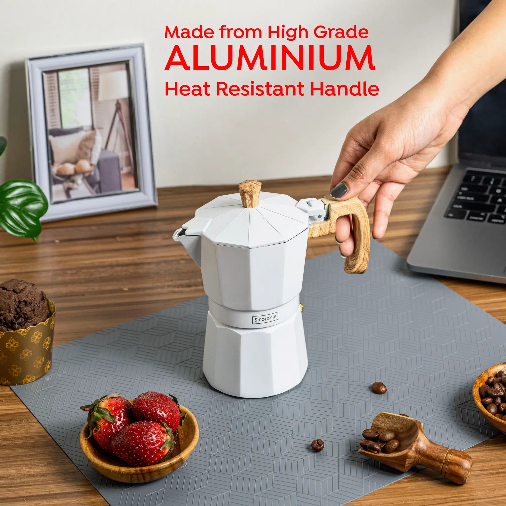 A white aluminium moka pot with a wooden handle and lid knob sits on a gray mat on a wooden table, with a hand holding the handle. Red text at the top reads 'Made from High Grade Aluminium Heat Resistant Handle'.