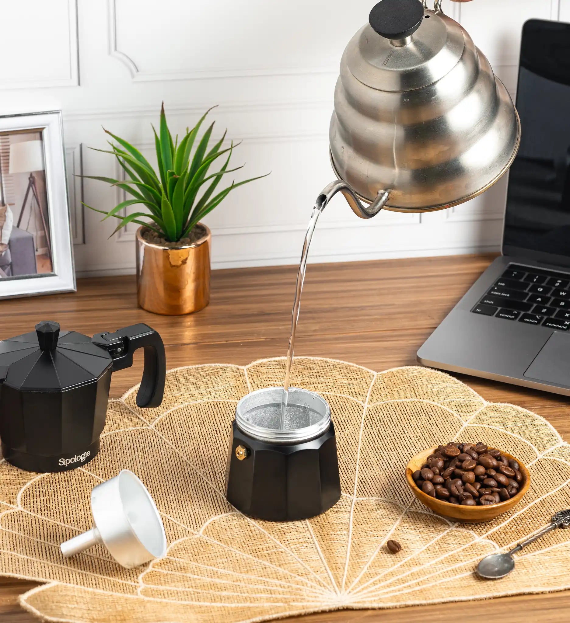 A black moka pot coffee maker sits on a woven placemat on a wooden desk, with hot water being poured into it from a stainless steel kettle. A second moka pot, a small bowl of coffee beans, a white cup, and a spoon are nearby, with a laptop and potted plant in the background.