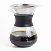 A glass pour-over coffee maker with a black silicone collar, clear glass body with measurement markings, and a stainless steel mesh filter cone, shown filled with coffee on a white background.