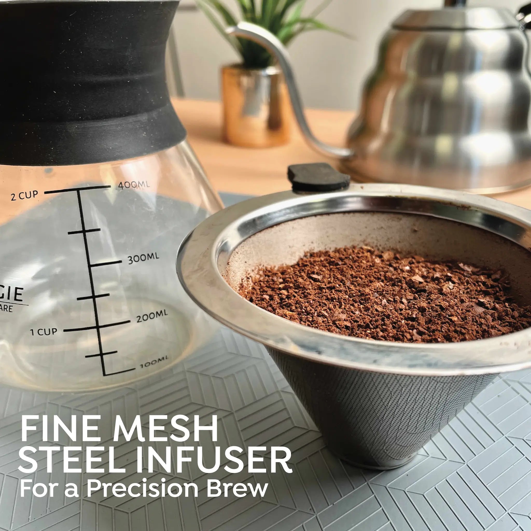 A metal mesh coffee infuser filled with ground coffee, shown beside a glass pour-over carafe with printed measurement markings and a stainless steel kettle in the background. Text at the bottom reads 'FINE MESH STEEL INFUSER For a Precision Brew'.