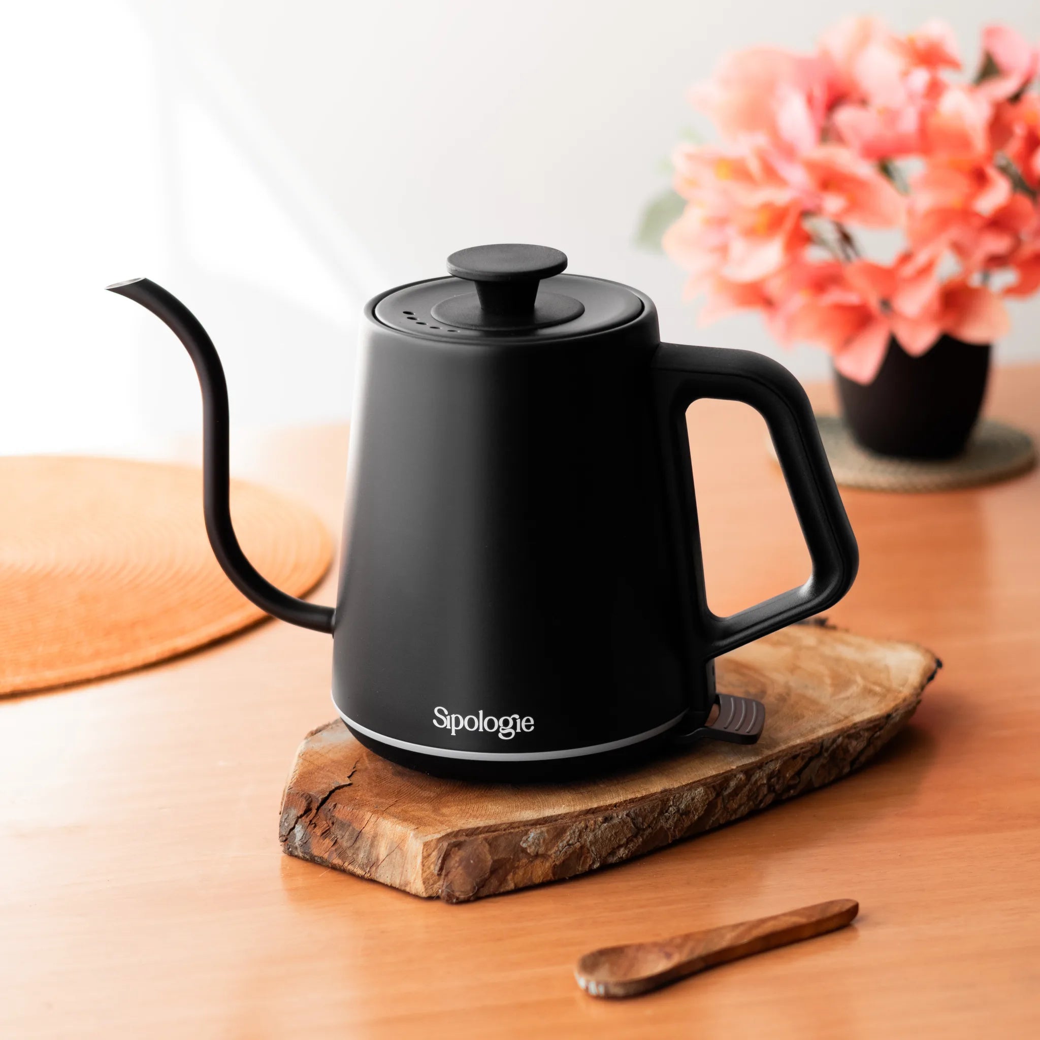 A matte black Spologie electric kettle with a long spout and integrated handle, resting on a rustic wooden slice coaster on a wooden table. A small wooden spoon is in the foreground, and a blurred vase of pink flowers is in the background.