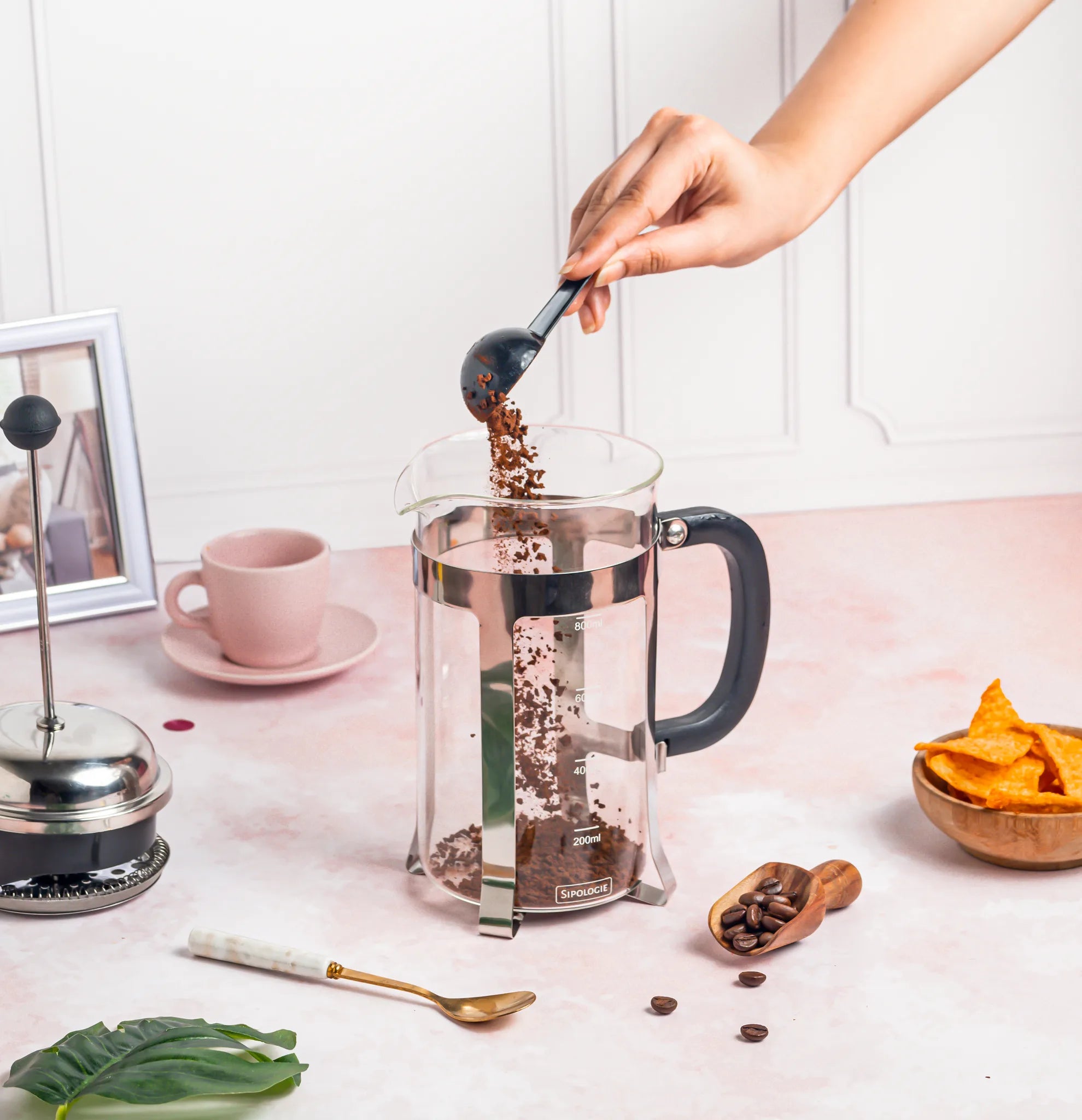 A clear glass French press with a black handle and metal collar sits on a pale pink tabletop. A hand is pouring coffee beans from a black spoon into the press.
