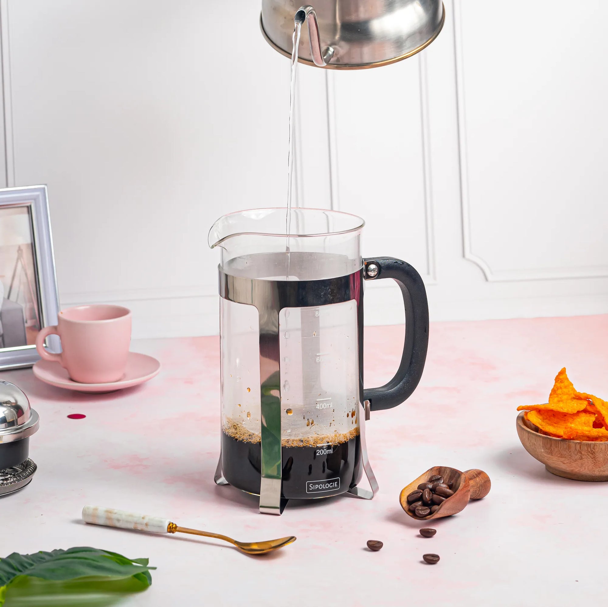A glass French press coffee maker with a black handle and silver metal components, shown with hot water being poured into it from a metal kettle. The press is set on a pink surface with coffee beans, a spoon, and other small kitchen props around it.