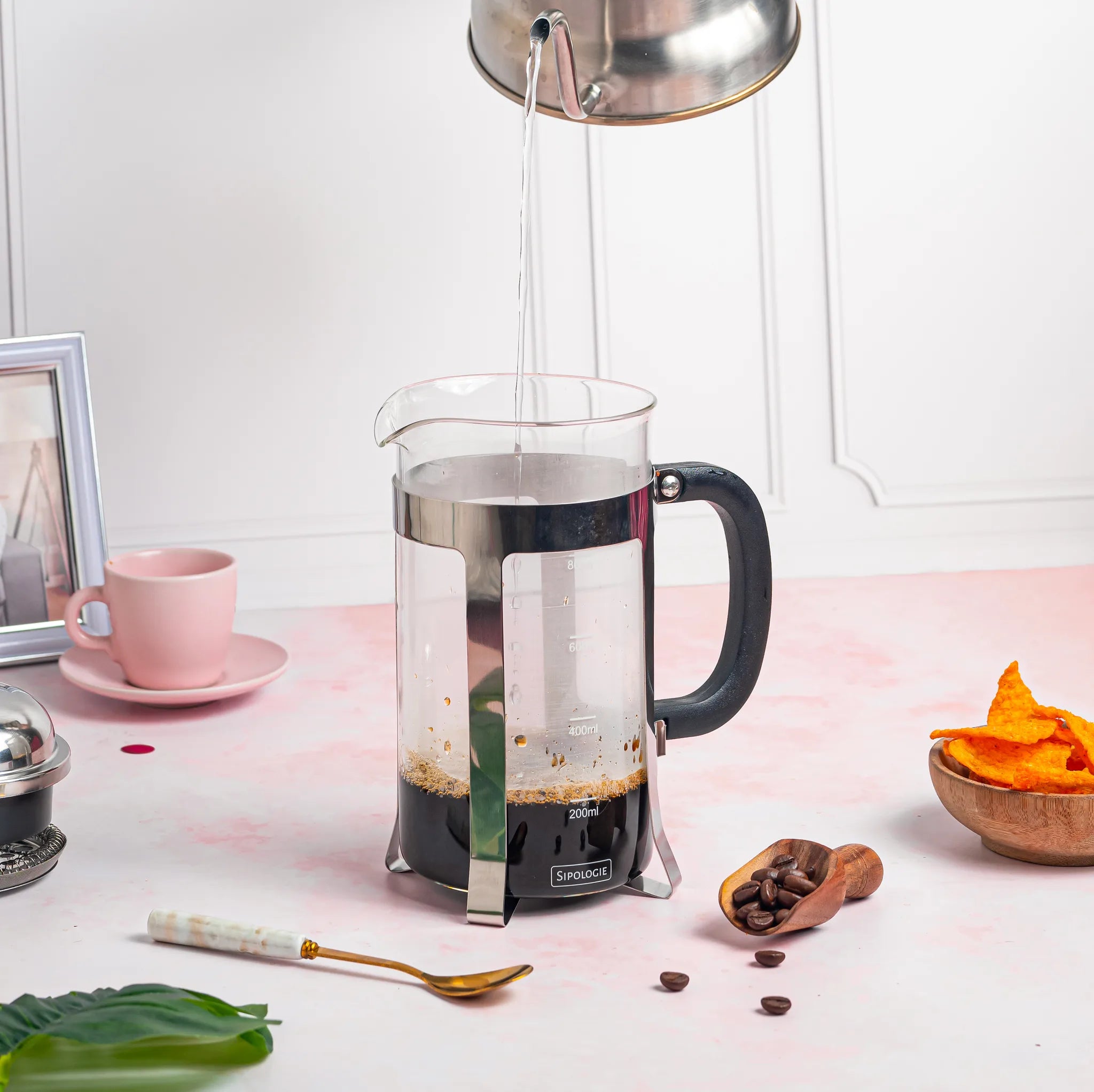 A glass French press coffee maker with a black handle and silver metal components, shown with hot water being poured into it from a metal kettle. The press is set on a pink surface with coffee beans, a spoon, and other small kitchen props around it.