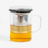 A clear glass teapot with a handle, a metal infuser, and a black lid, filled with amber tea, shown on a light gray background.