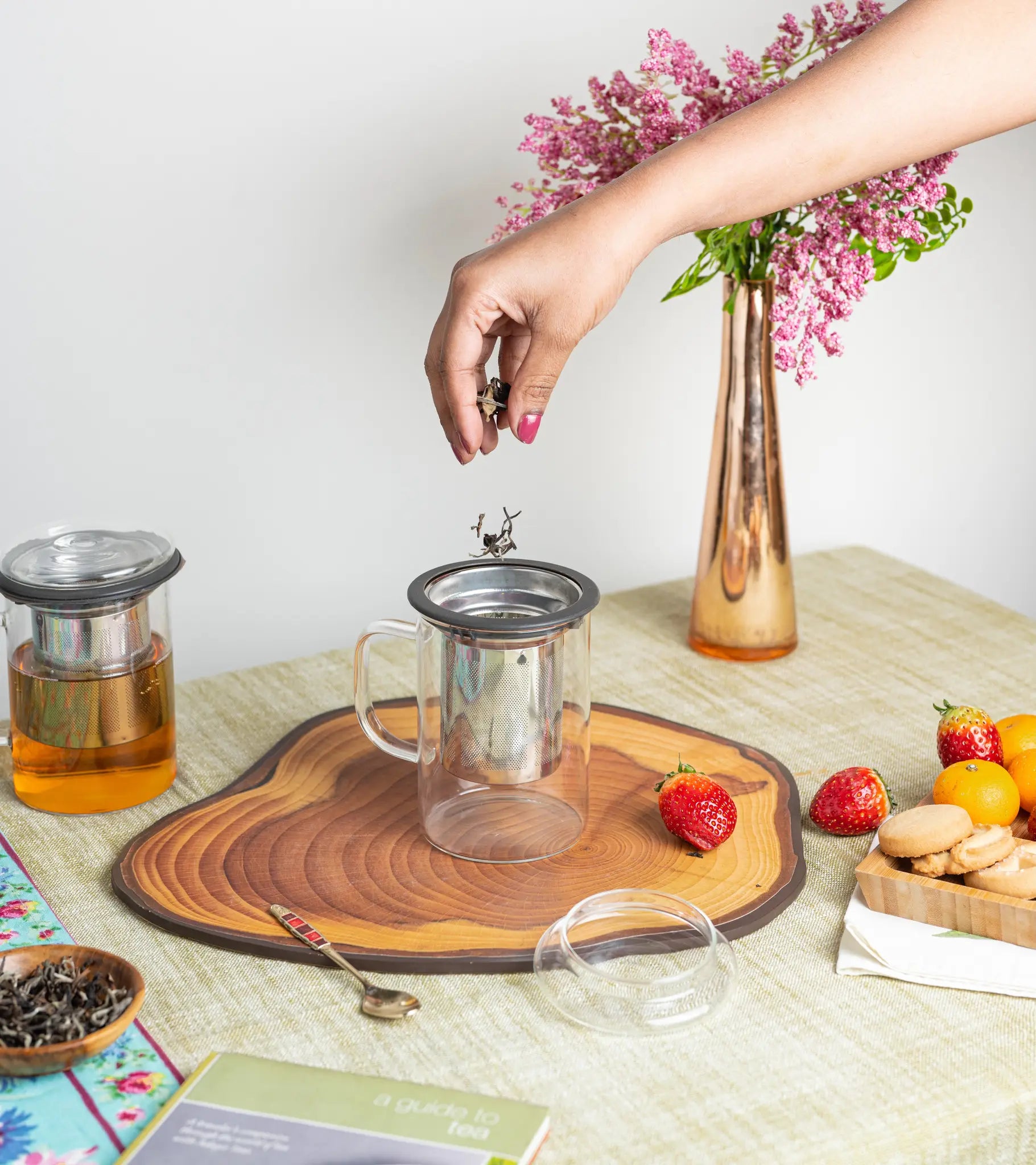 A clear glass teapot with a stainless steel infuser and lid is shown on a table, with a hand placing the lid onto the infuser. The scene includes a copper vase with pink flowers, strawberries, cookies, and another glass jar with amber liquid.