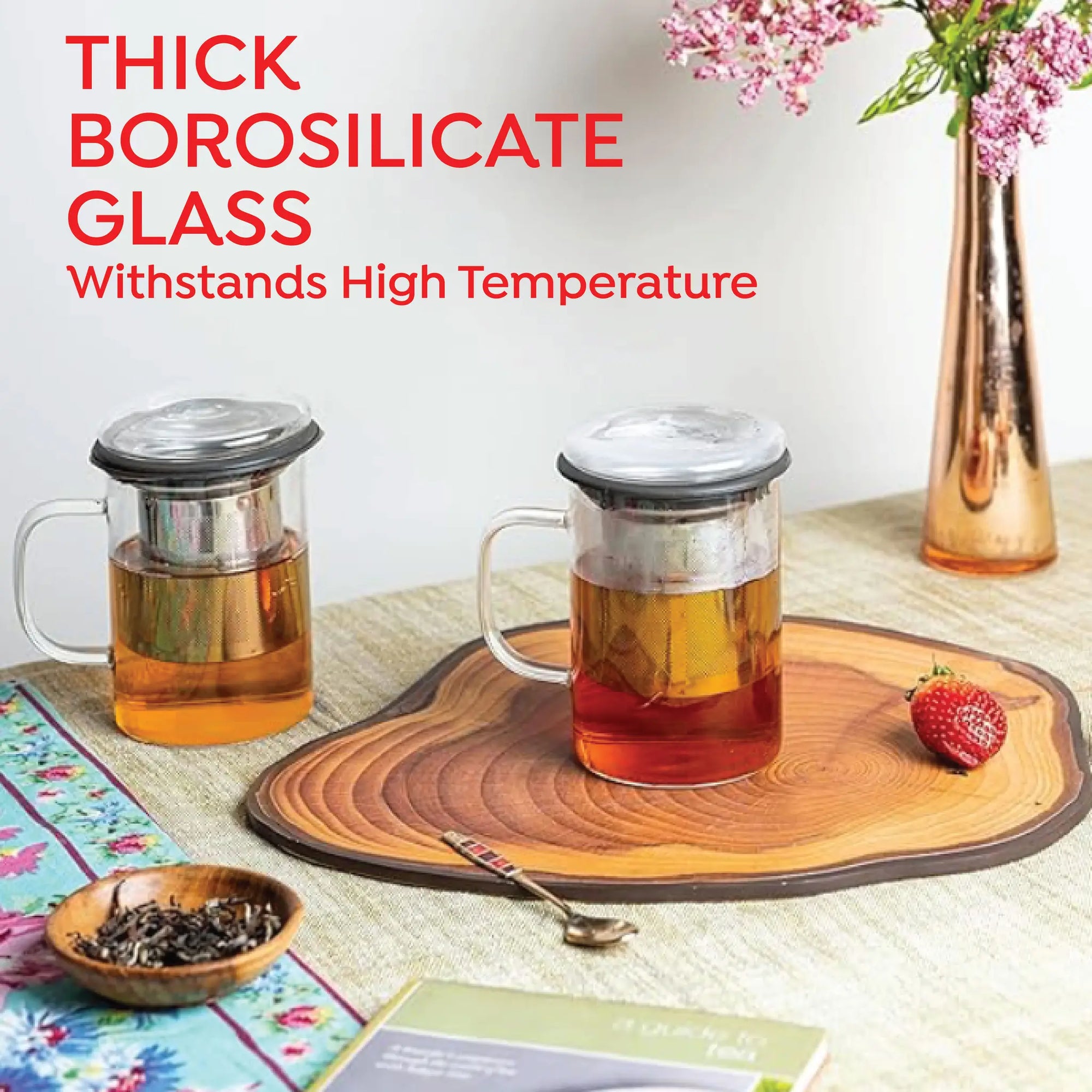 Two thick borosilicate glass teapots with metal infusers and lids are shown on a tabletop with tea, a strawberry, a wooden slice board, and decorative props. Large text at the top reads 'THICK BOROSILICATE GLASS Withstands High Temperature'.