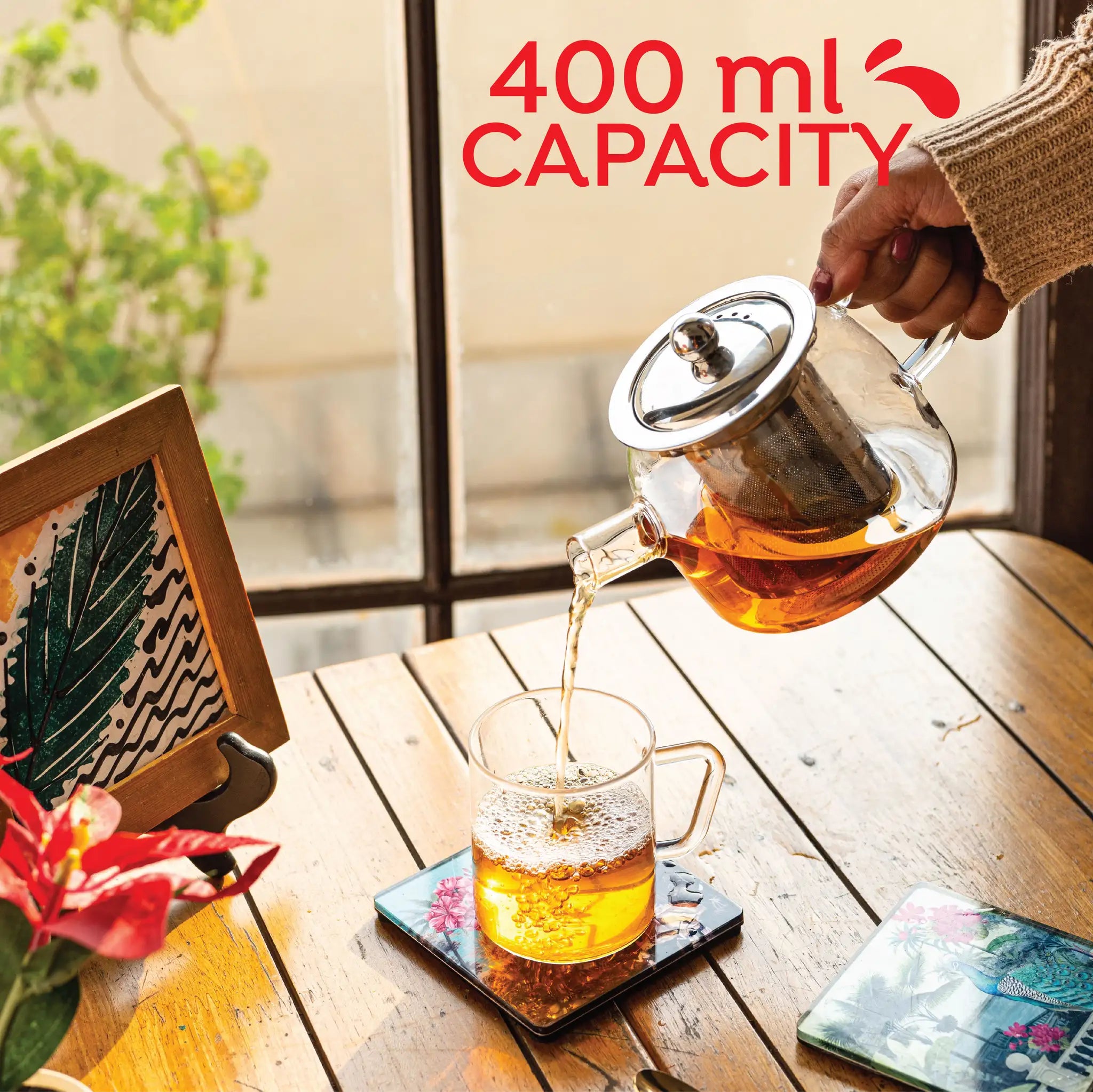 A clear glass teapot with a metal infuser strainer is pouring amber tea into a matching clear glass mug on a wooden table. A hand holds the teapot, and the text '400 ml CAPACITY' appears in the top right.