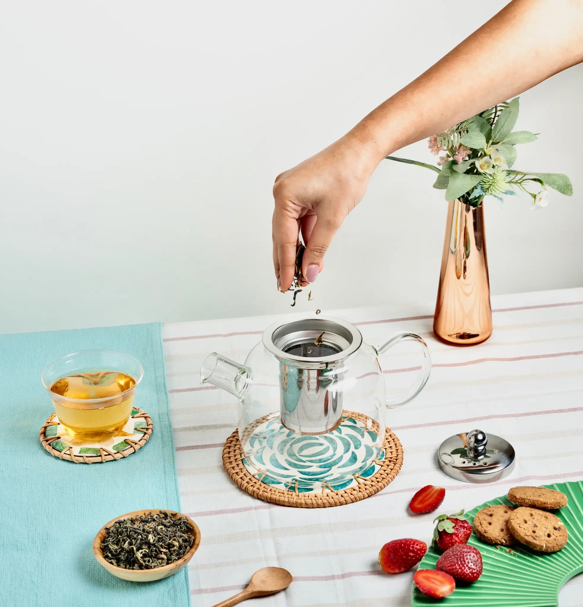 A clear glass teapot with a stainless steel infuser and lid is shown on a table, with a hand lifting the lid. The scene includes a glass cup of tea, loose leaves, strawberries, cookies, a woven coaster, and a copper vase with flowers.