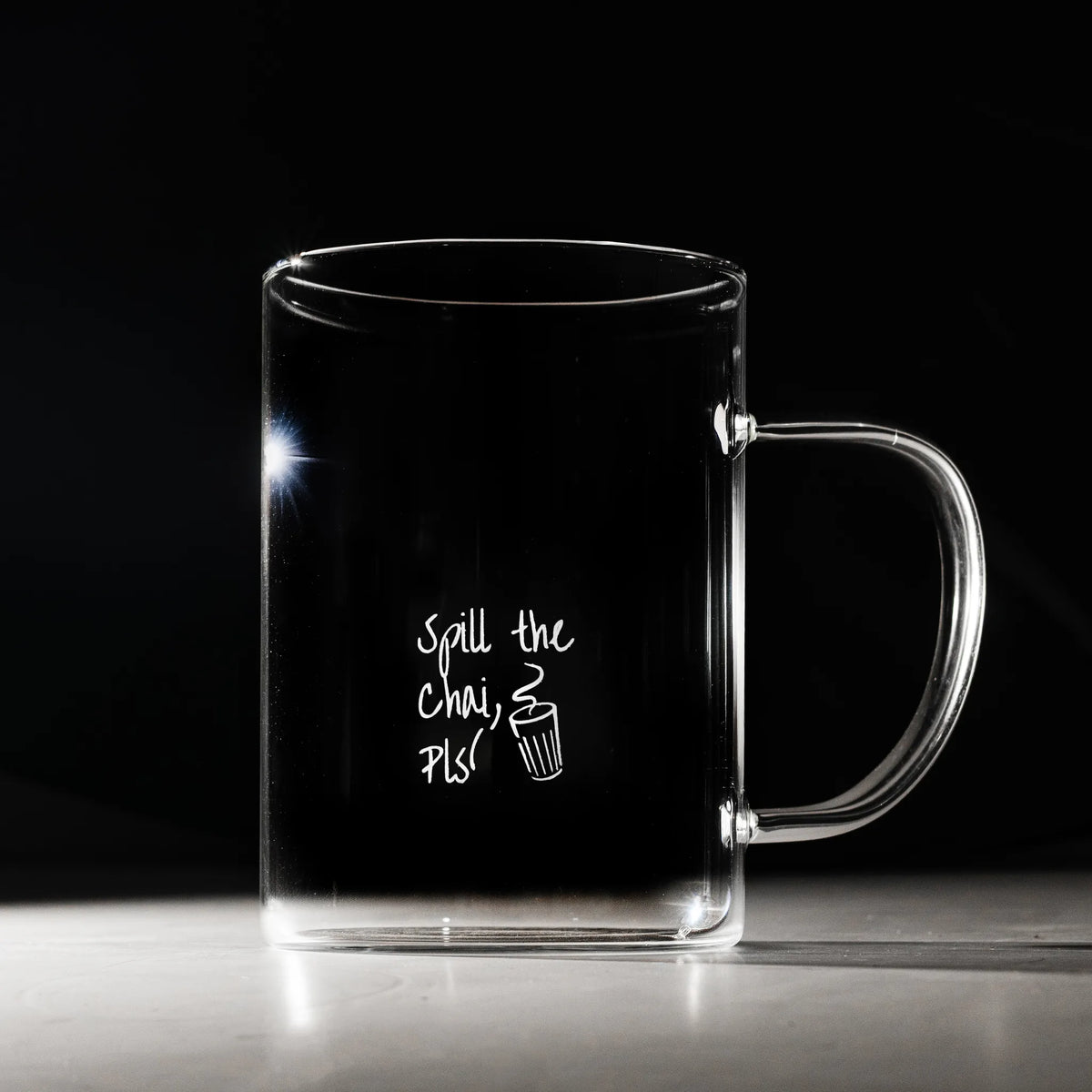 A clear glass mug with a handle, featuring white printed text that reads 'Spill the chai, pls' alongside a small chai cup illustration, set against a dark background.