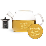 A clear glass teapot filled with amber tea, shown on a white background with a small black infuser and a circular printed graphic overlay. The glass features the text 'me, myself, and chai!' in yellow with a chai cup icon.