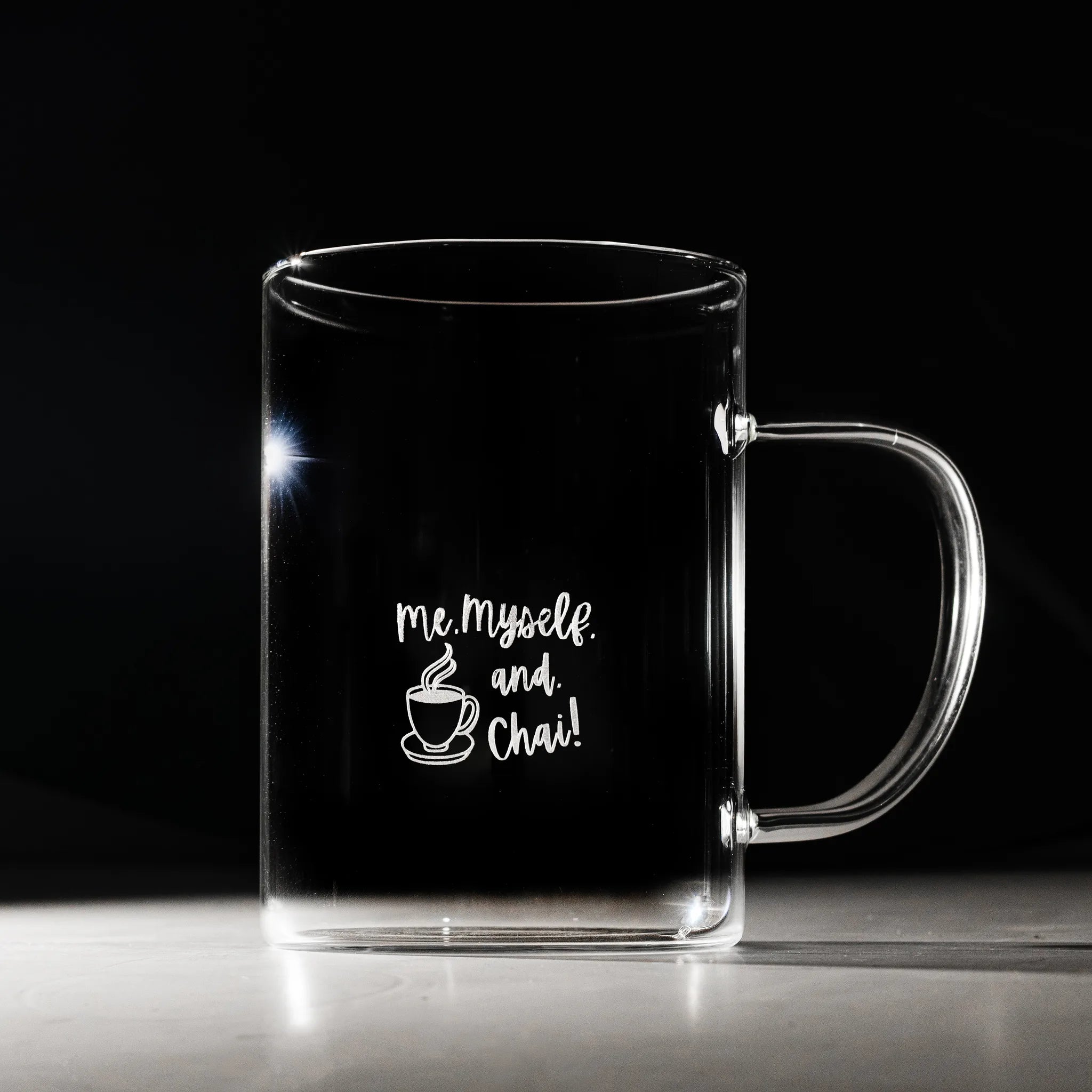 A clear glass mug with a handle, featuring the white printed text 'me, myself, and Chai!' with a small cup graphic, set against a dark background.