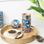 A decorative tea tin with a blue, white, and red floral-and-bird pattern, shown on a round wooden tray with loose tea, a small bowl, and a metal scoop. A matching patterned lid sits beside the tin, with green leaves entering from the top right.