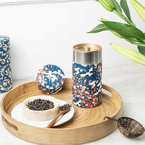 A decorative tea tin with a blue, white, and red floral-and-bird pattern, shown on a round wooden tray with loose tea, a small bowl, and a metal scoop. A matching patterned lid sits beside the tin, with green leaves entering from the top right.
