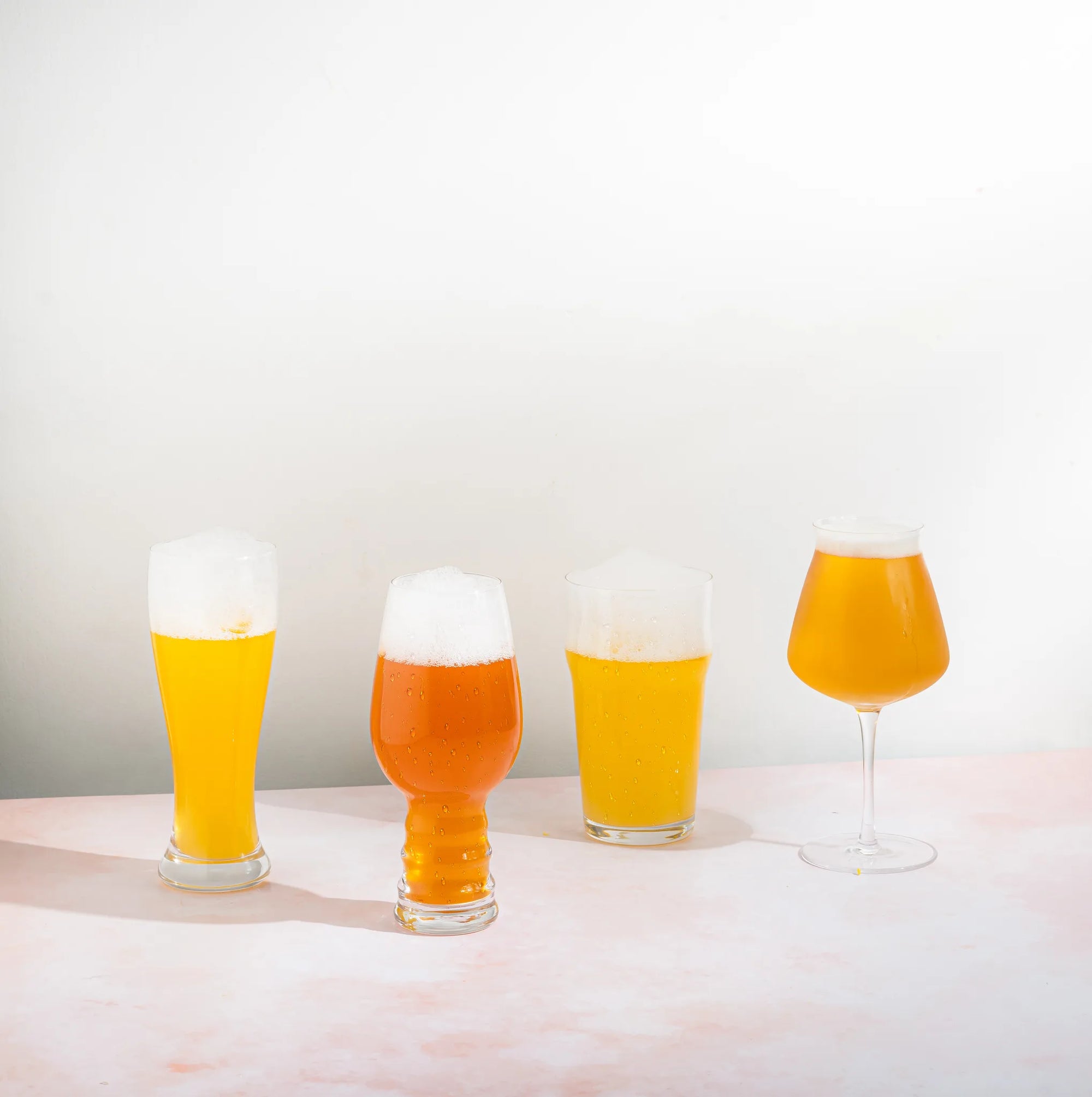 Four beer glasses of different shapes and sizes filled with golden beer and topped with white foam, arranged on a pale pink surface against a white background.