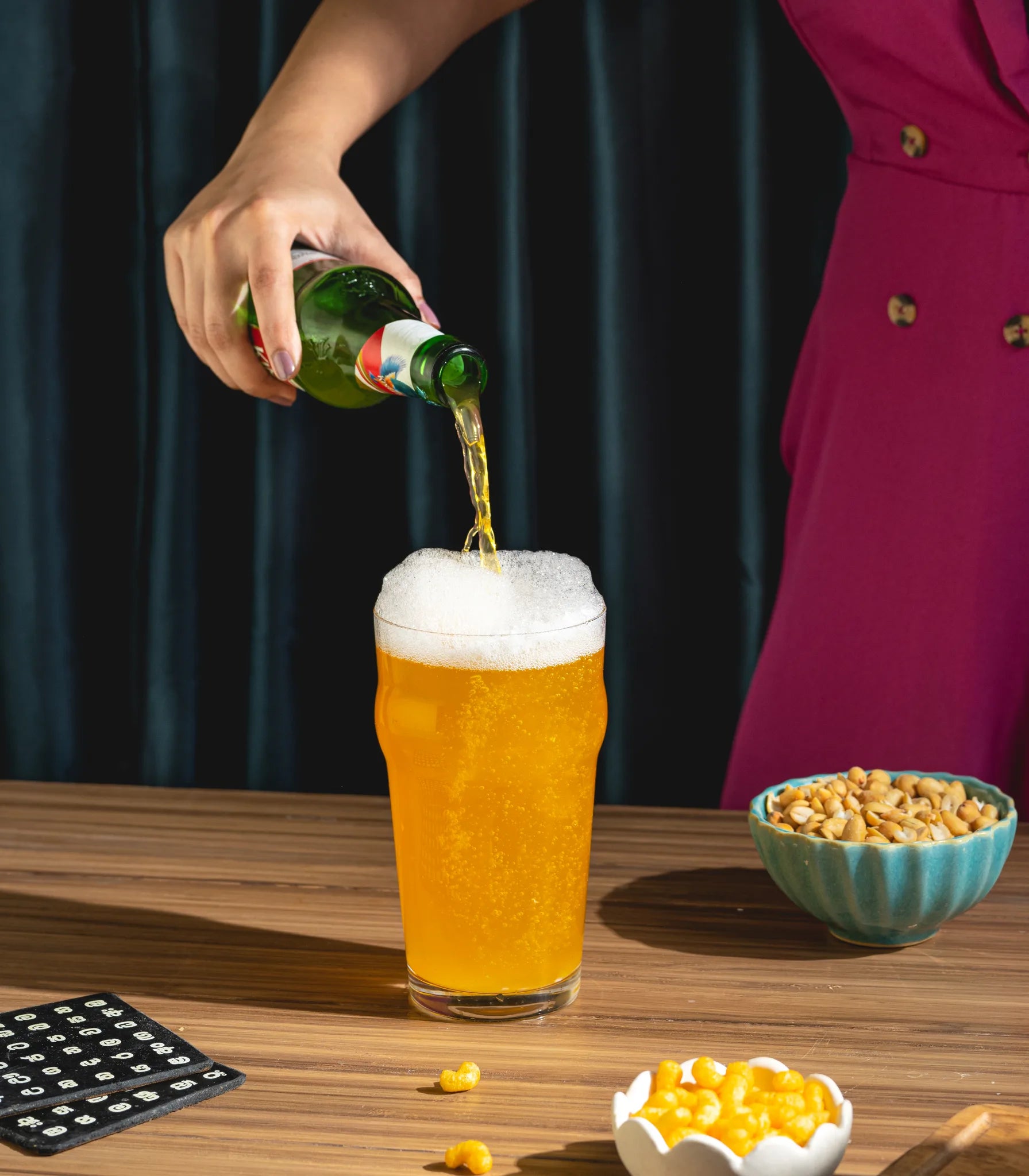 A hand pours golden beer from a green glass bottle into a tall glass of foamy lager on a wooden table, with a bowl of peanuts and a person in a magenta dress partially visible in the background.