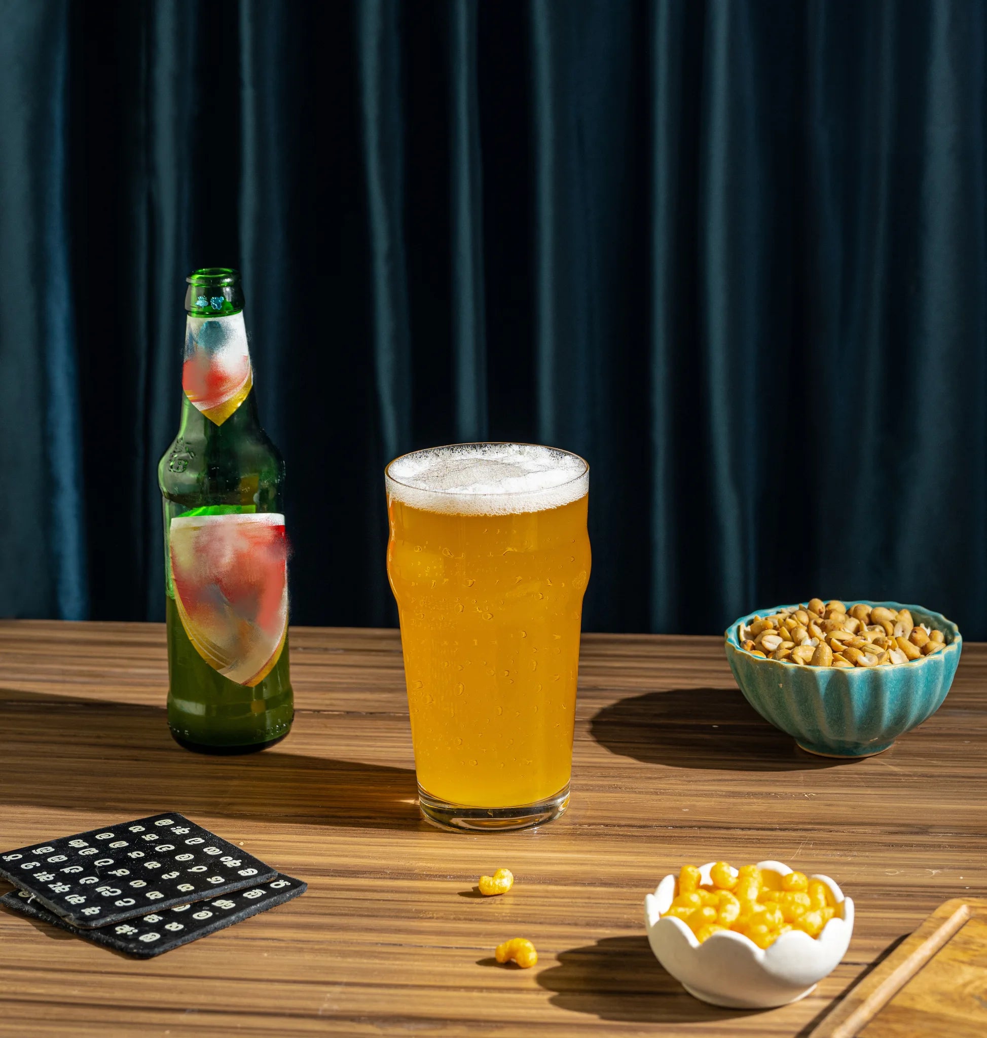 A glass of golden beer with white foam sits on a wooden table alongside a green beer bottle, bowls of popcorn kernels, and a black popcorn tray against a dark blue curtain background.