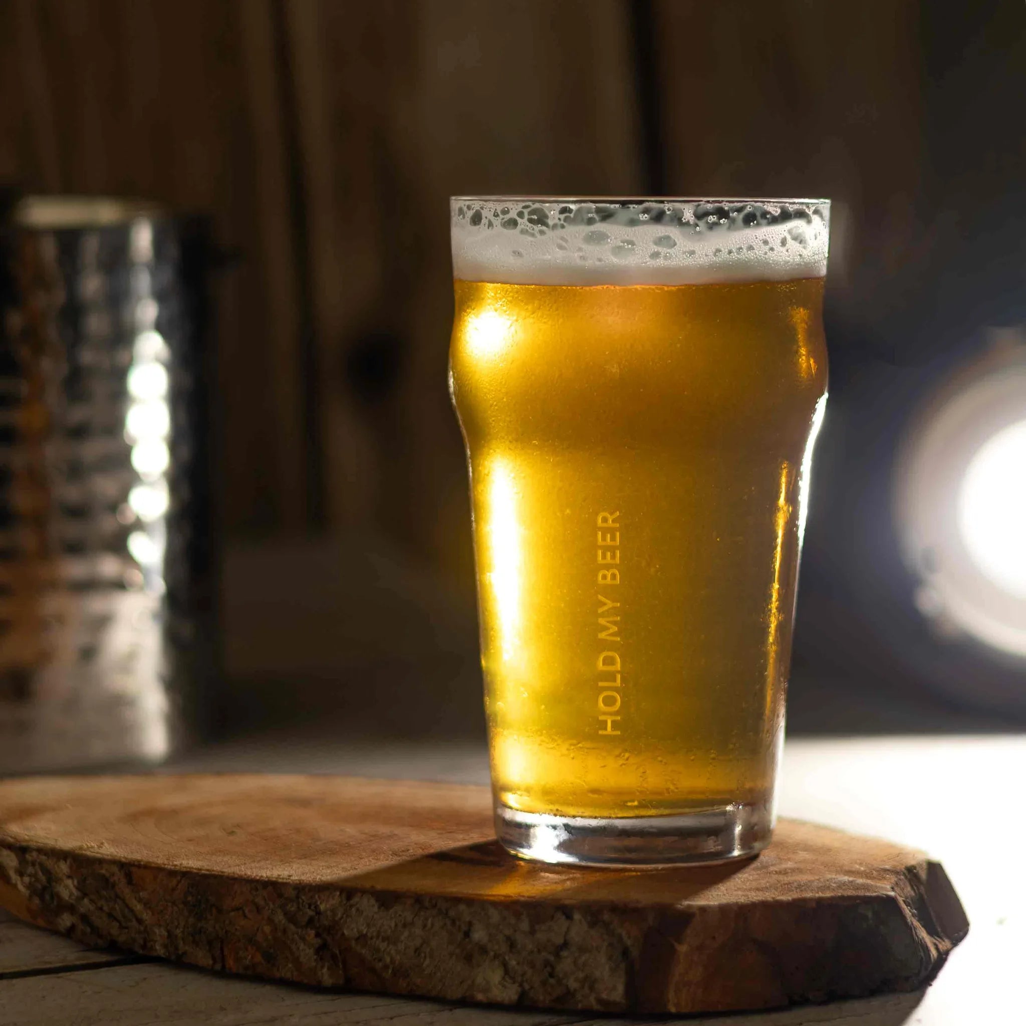 A clear pint glass filled with golden beer, standing on a rustic wooden slice with a blurred metal container in the background.
