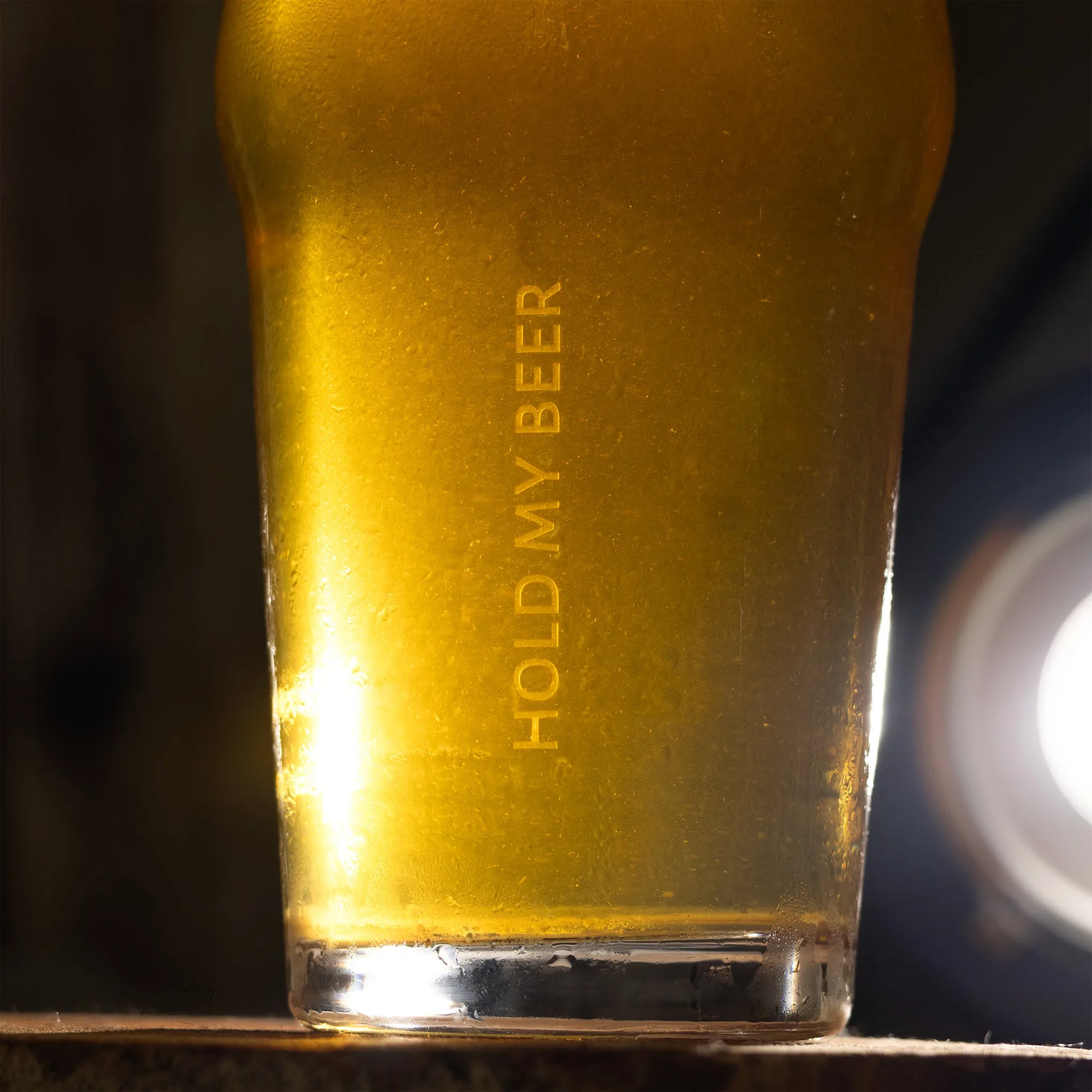 A close-up of a pint glass filled with golden beer, featuring the text 'HOLD MY BEER' printed vertically on the side, set against a dark background with a bright light flare on the right.