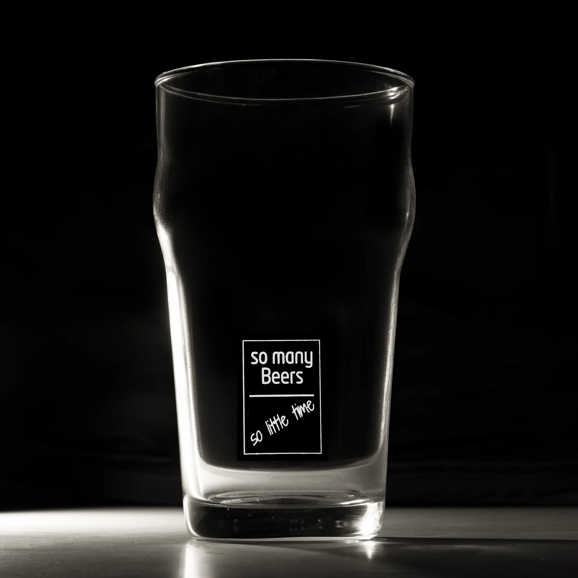 A clear pint glass with a black printed graphic that reads 'so many Beers so little time' on a dark background.