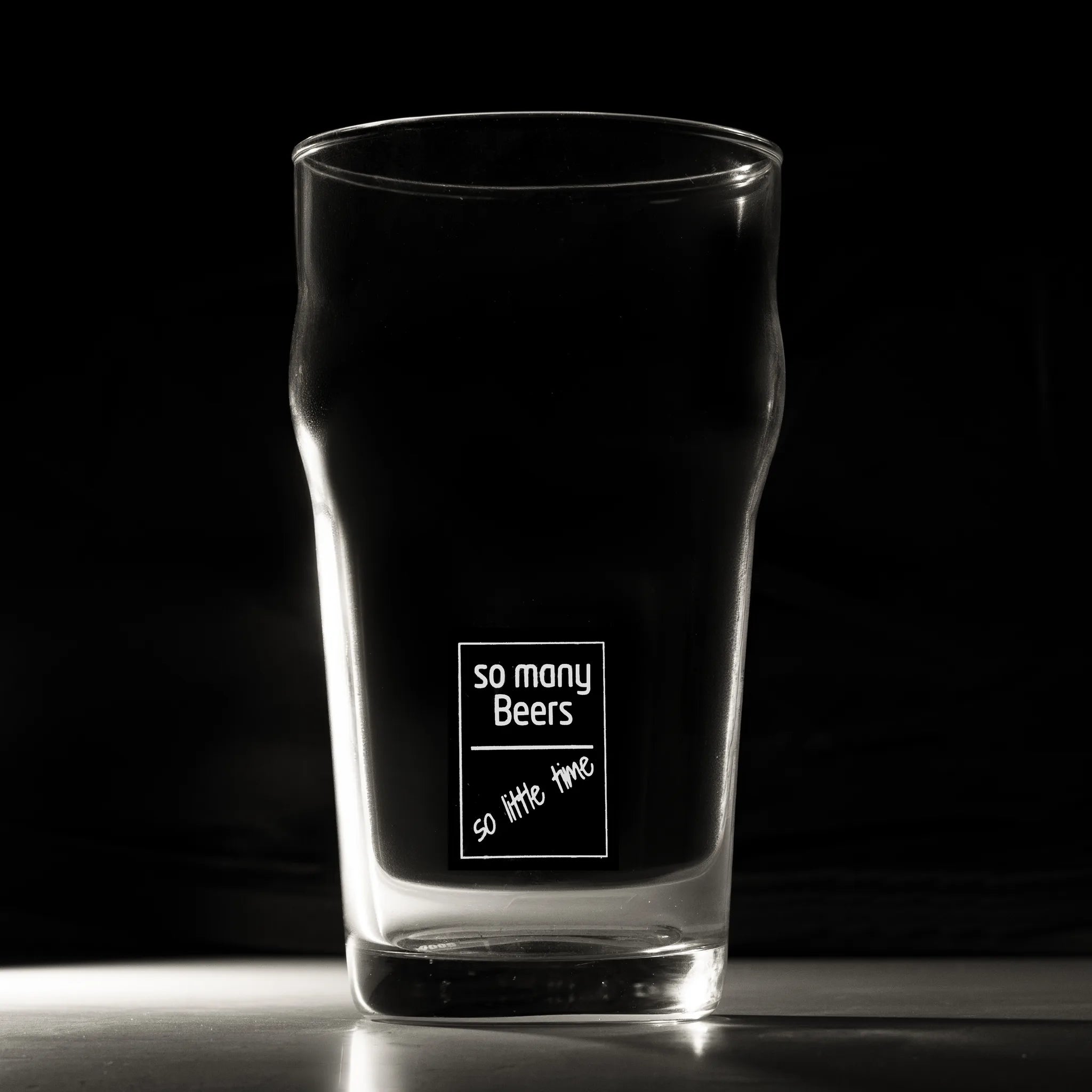 A clear pint glass with a black printed graphic that reads 'so many Beers so little time' on a dark background.