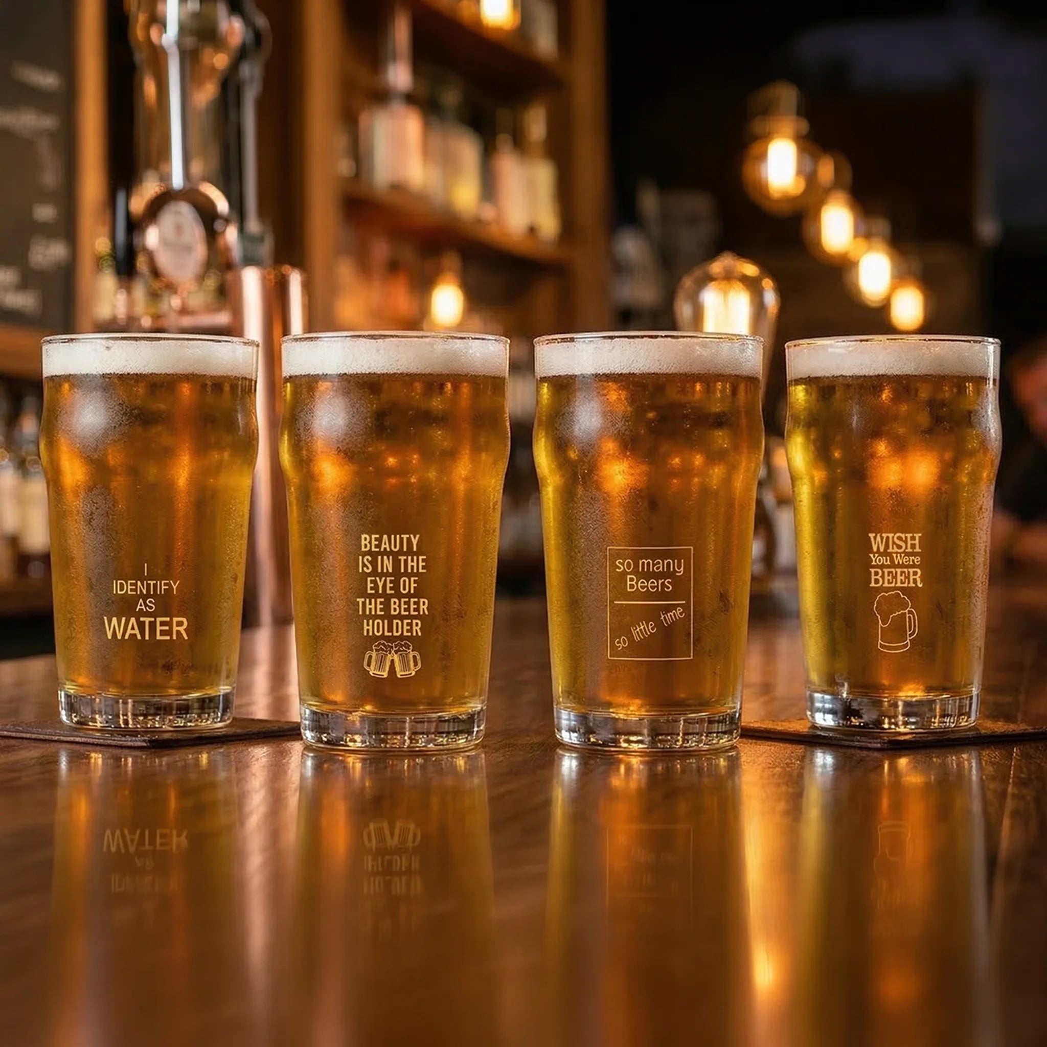 Four pint glasses filled with golden beer lined up on a wooden bar counter in a dimly lit pub. Each glass has a different frosted etched quote on the front, including 'I IDENTIFY AS WATER', 'BEAUTY IS IN THE EYE OF THE BEER HOLDER', 'so many Beers', and 'WISH FOR MORE BEER'.