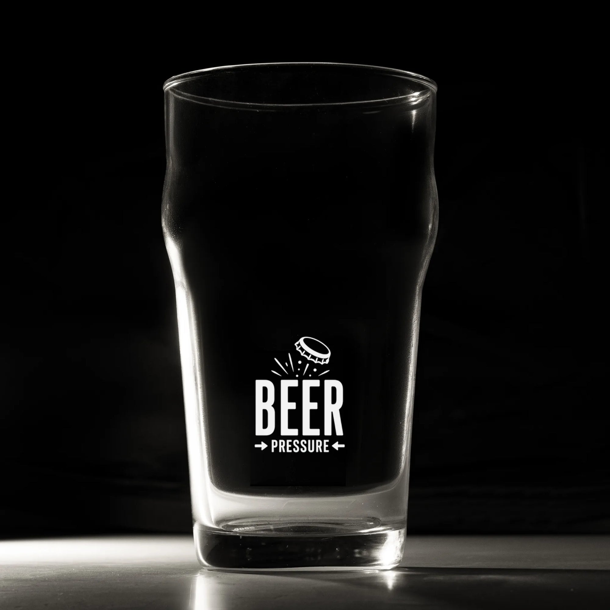 A clear pint glass with a black printed graphic that reads 'BEER PRESSURE' and a small beer bottle illustration, photographed against a dark studio background.