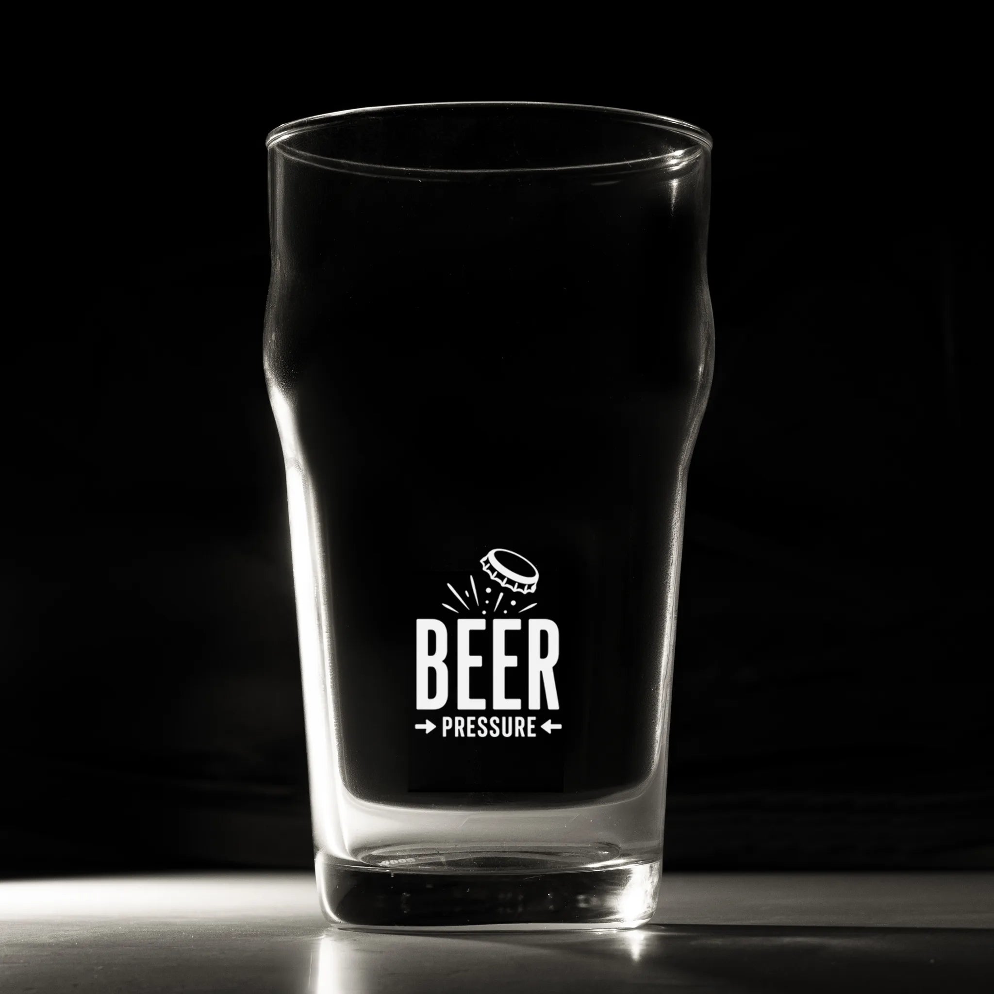 A clear pint glass with a black printed graphic that reads 'BEER PRESSURE' and a small beer bottle illustration, photographed against a dark studio background.