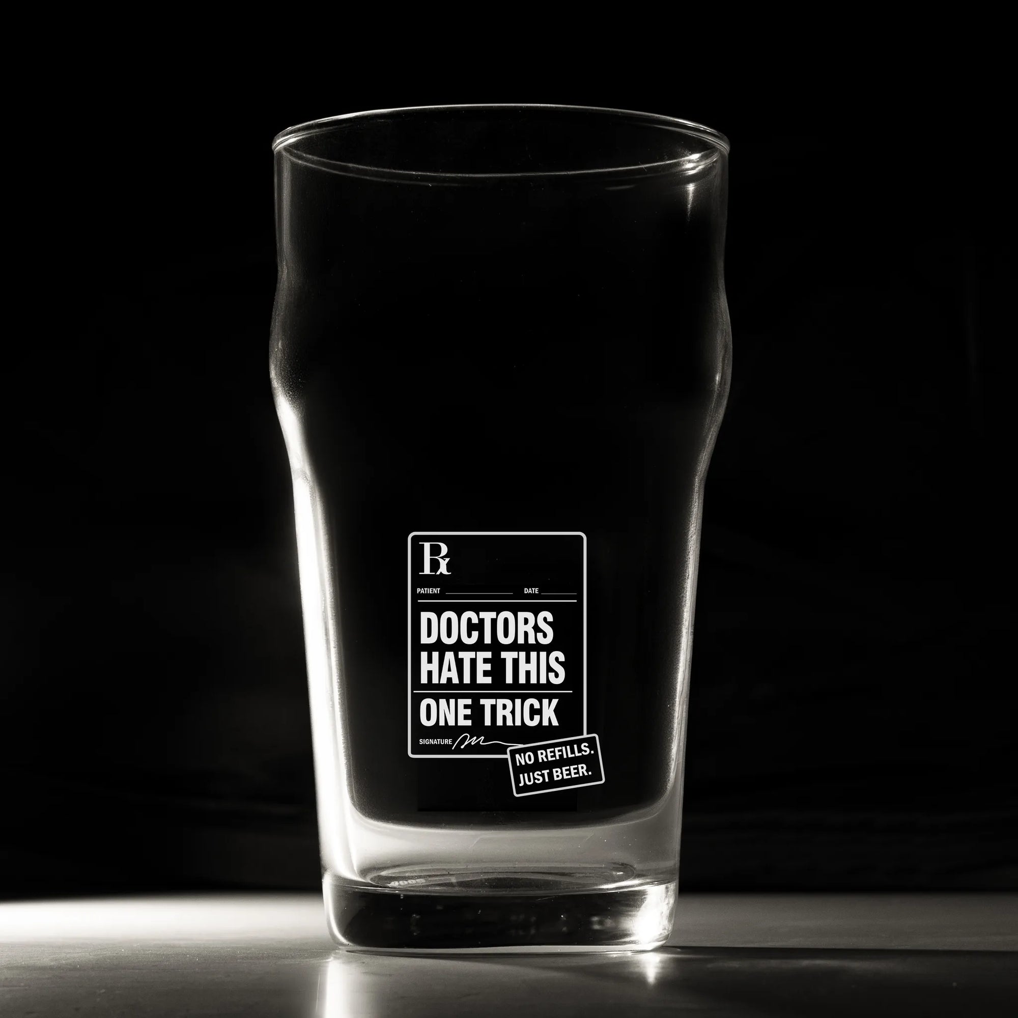 A clear pint glass with a black printed graphic that reads 'DOCTORS HATE THIS ONE TRICK' and a small badge reading 'NO REFILL - JUST REBORN.' The glass is photographed on a dark studio background with a reflective surface below.