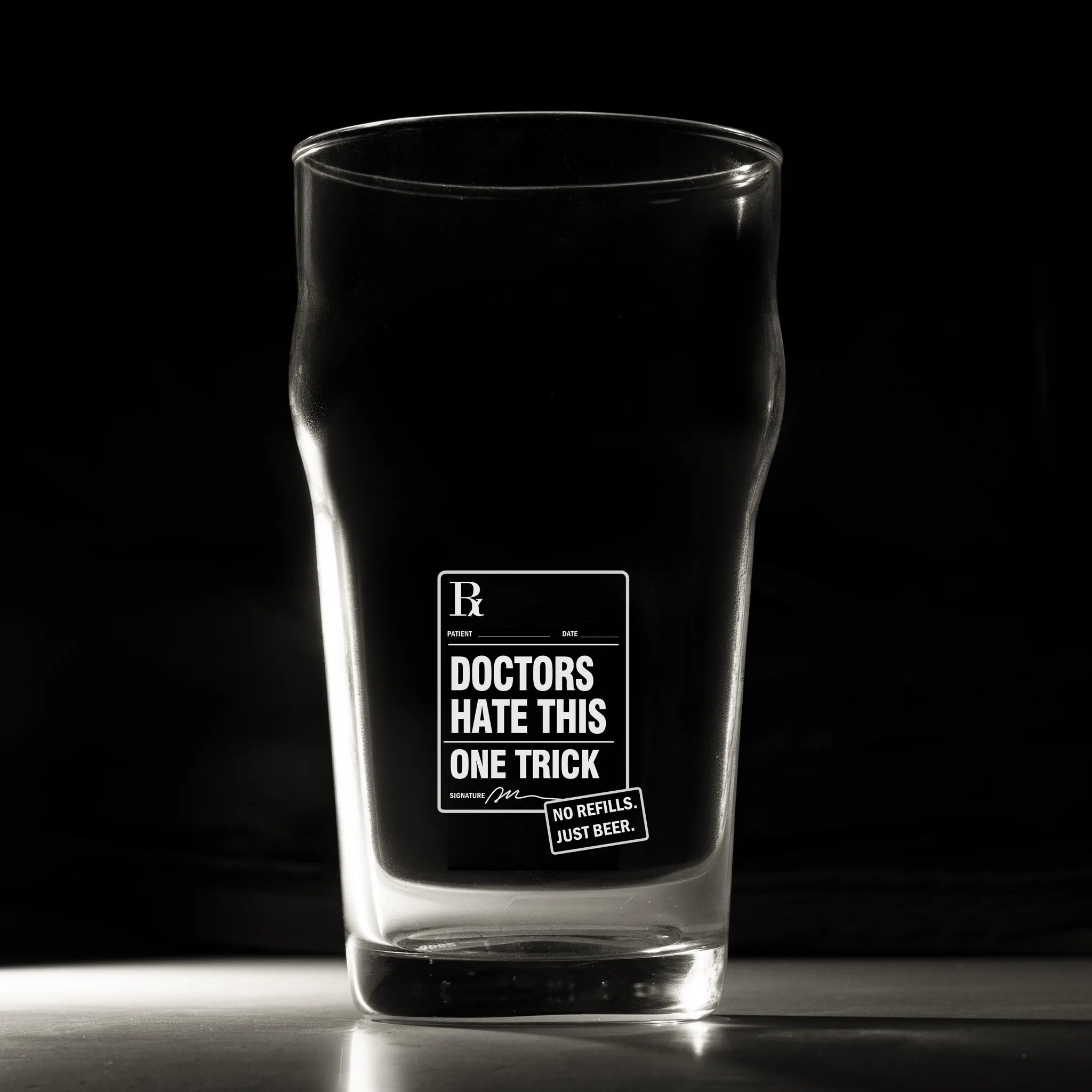 A clear pint glass with a black printed graphic that reads 'DOCTORS HATE THIS ONE TRICK' and a small badge reading 'NO REFILL - JUST REBORN.' The glass is photographed on a dark studio background with a reflective surface below.